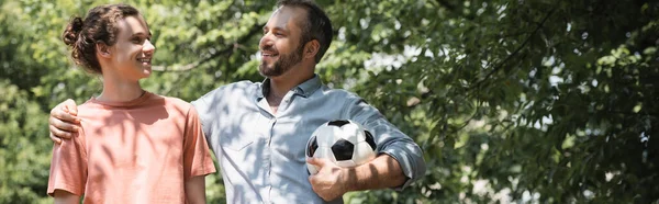 Wesoły Mężczyzna Trzyma Piłkę Nożną Podczas Przytulania Szczęśliwy Nastoletni Syn — Zdjęcie stockowe