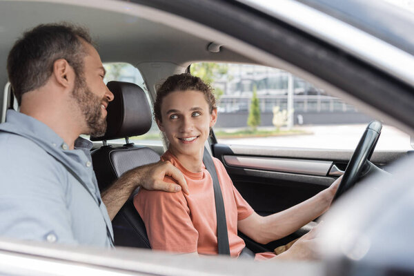cheerful father putting hand on shoulder of teenage son while teaching him how to drive car 