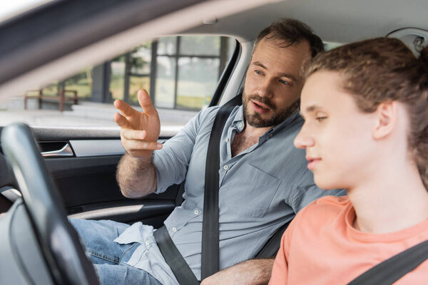 bearded dad gesturing while explaining to teenage son how to drive car 