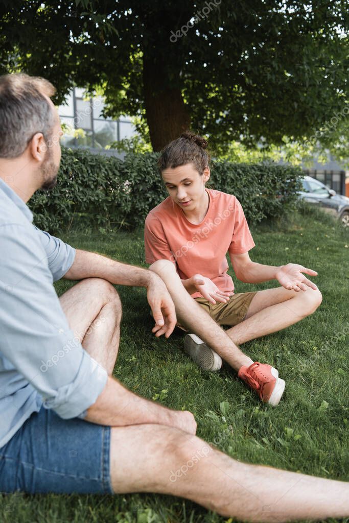 Confused teenage boy sitting with crossed legs next to father on green park
