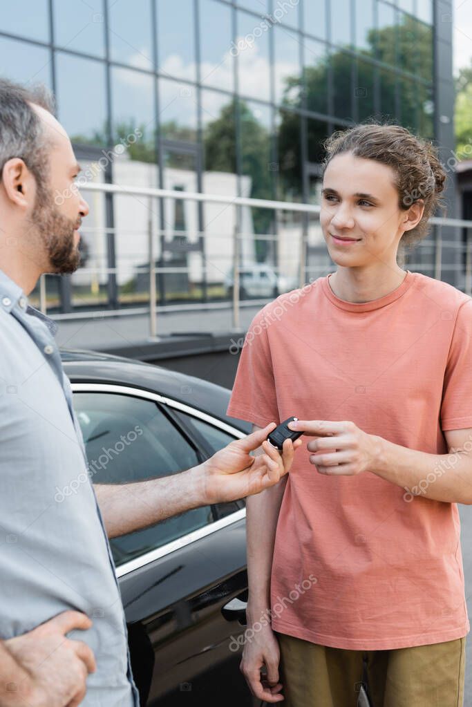 Happy bearded man giving car key to cheerful teenage son near automobile
