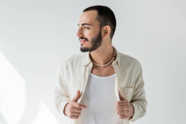 Smiling and bearded gay man in casual clothes and pearl necklace touching shirt and looking away while posing on grey background with sunlight  clipart