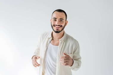 Gri arka planda poz verirken kameraya bakan bej gömlekli ve inci kolyeli neşeli ve sakallı genç homoseksüel adam.