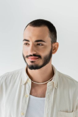 Portrait of young bearded homosexual man in beige shirt, sleeveless t-shirt and pearl necklace looking away while standing isolated on grey  clipart