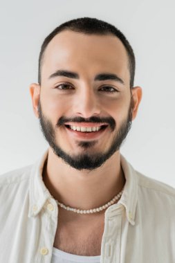 Portrait of young and happy homosexual man in casual clothes and pearl necklace looking at camera while standing and smiling isolated on grey  clipart