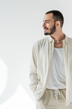 Sakallı ve kısa saçlı, bej gömlekli ve şık inci kolyeli eşcinsel adam gözlerini kaçırıyor ve gri arka planda, güneş ışığıyla birlikte doğal kumaşlardan yapılmış pantolonun cebinde el ele tutuşuyor. 