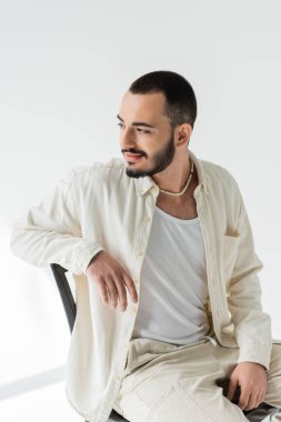 Esmer, bej elbiseli, doğal kumaşlı ve inci kolyeli genç bir homoseksüel. Gri arka planda, güneş ışığı olan bir sandalyede otururken rahatlıyor. 