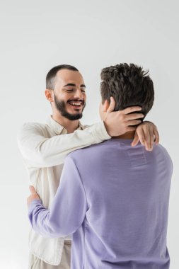 Olumlu sakallı eşcinsel bir adam, üstü kapalı gömlekli, boynuna dokunan, esmer erkek arkadaşına sarılan ve gri renkli bir elbisenin üzerinde tek başına duran. 