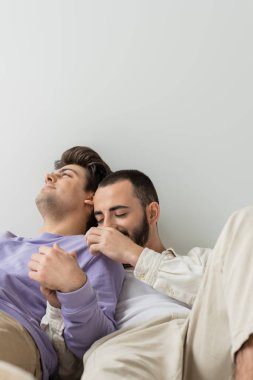Genç homoseksüel adam omuzlarına dokunuyor ve gri arka planda birlikte otururken esmer erkek arkadaşının elini tutuyor.