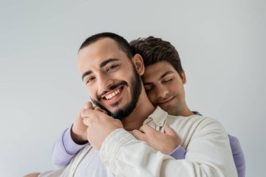 Portrait of positive and bearded gay man holding hand of young brunette boyfriend near face and looking at camera isolated on grey  clipart