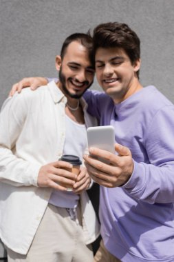 Bulanık eşcinsel adam, güler yüzlü erkek arkadaşına sarılıp kahve içiyor ve gri binanın yanındaki cep telefonunu kullanıyor. 