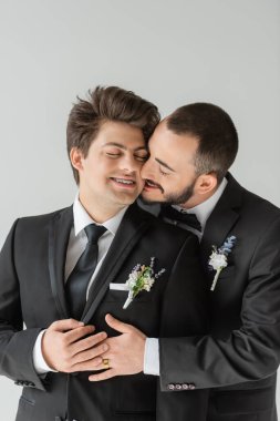 Genç ve sakallı eşcinsel damat, yaka çiçeğiyle sarılıp öpüşen, gözleri kapalı bir şekilde gülümseyen bir erkek arkadaş.  