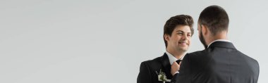 Sakallı eşcinsel adam, gri pankartta izole edilmiş nikah töreninde zarif ve klasik bir takım elbise ve çiçek yaka çiçekli diş telleriyle gülen damat kravatını düzeltiyor. 