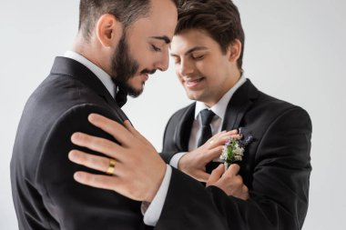 Olumlu ve sakallı eşcinsel damat, gri renkte izole edilmiş düğün töreninde diş teli takmış şık erkek arkadaş çiçeğini düzeltiyor.  