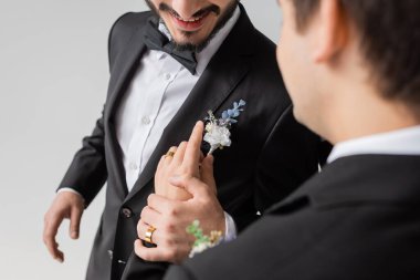Gri renkte izole edilmiş düğün kutlaması sırasında gülümseyen erkek arkadaş elbisesine butonye takan bulanık homoseksüel damat manzarası. 