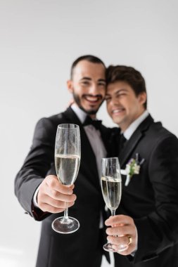 Blurred lgbt couple in elegant formal wear smiling and showing champagne glasses at camera during wedding celebration isolated on grey  clipart