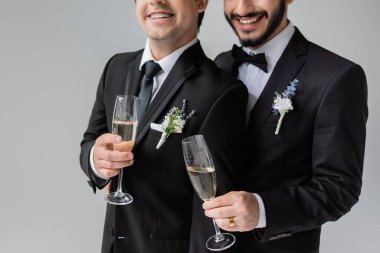 Cropped view of smiling homosexual grooms in classic attire with floral boutonnieres holding glasses of champagne while celebrating wedding isolated on grey  clipart