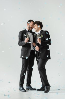Klasik takım elbiseli, pozitif, aynı cinsiyetten çiftler ellerinde şampanya bardakları tutarken gri arka planda düşen konfeti altında düğünü kutluyorlar.
