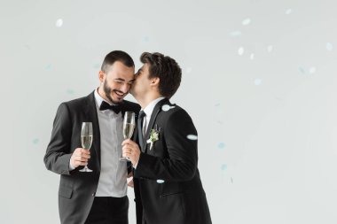 Resmi olarak genç homoseksüel damat, gri arka planda düşen konfeti altında şampanya tutan sakallı bir erkek arkadaş giyer.