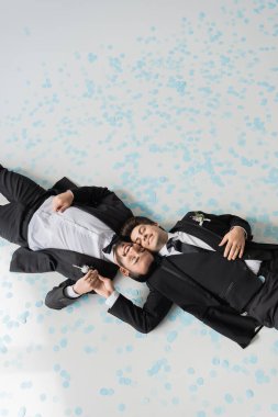 Top view of young and smiling homosexual grooms with closed eyes in suits holding hands while lying together on festive confetti on grey background  clipart