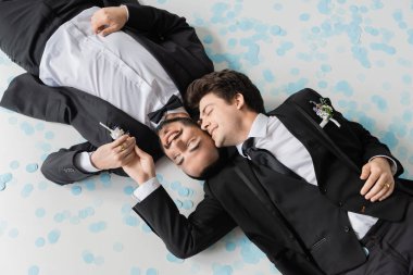 Gülümseyen eşcinsel damadın, klasik takım elbiseli, sakallı erkek arkadaşının ellerine dokunup gri arka plandaki düğün kutlaması sırasında şenlikli konfetide yan yana yatması. 