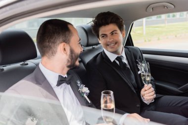 Smiling gay groom in formal wear holding glass of champagne and looking at boyfriend while sitting on backseat of car during honeymoon trip  clipart