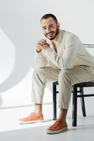 Полная длина улыбающийся коротковолосый гомосексуалист в бежевой повседневной одежде из натуральных тканей, сидящих на стуле и смотрящих в камеру на сером фоне со светом 