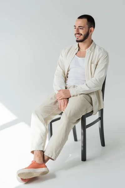 Carefree homosexual man in pearl necklace and casual clothes made of natural fabrics looking away while sitting on chair on grey background with sunlight