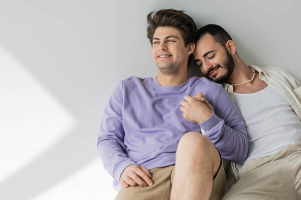 Smiling young gay man in casual clothes holding hand of bearded boyfriend with closed eyes while sitting together on grey background with sunlight 