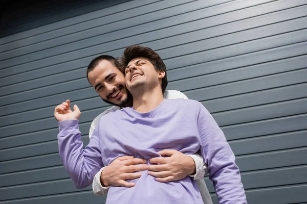 Low angle view of positive and bearded gay man hugging brunette boyfriend in sweatshirt and braces while looking at camera near building on urban street