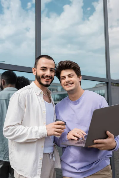 stock image Carefree same sex partners in casual clothes looking at camera while holding coffee to go and using laptop and standing near building on urban street 