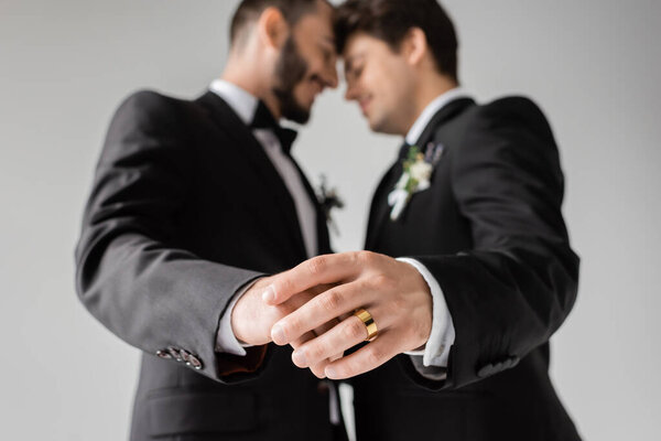 Side view of blurred same sex couple in formal wear with boutonnieres touching hands of each other with golden ring during wedding isolated on grey 