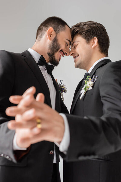 Low angle view of cheerful gay couple in formal wear with floral boutonnieres holding blurred hands during wedding isolated on grey 