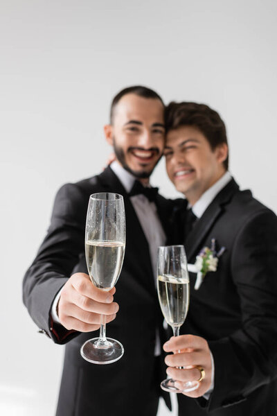 Blurred lgbt couple in elegant formal wear smiling and showing champagne glasses at camera during wedding celebration isolated on grey 