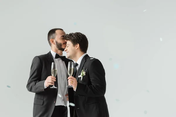 在婚礼上 有胡子的男同性恋新郎身穿正式婚纱 手持香槟酒 在灰色背景的彩礼下亲吻着年轻的男朋友 — 图库照片