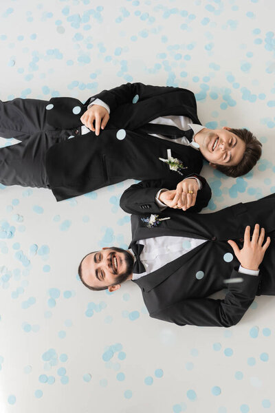 Top view of cheerful gay grooms in classis attire holding hands and looking at camera while lying on blue confetti during wedding on grey background 