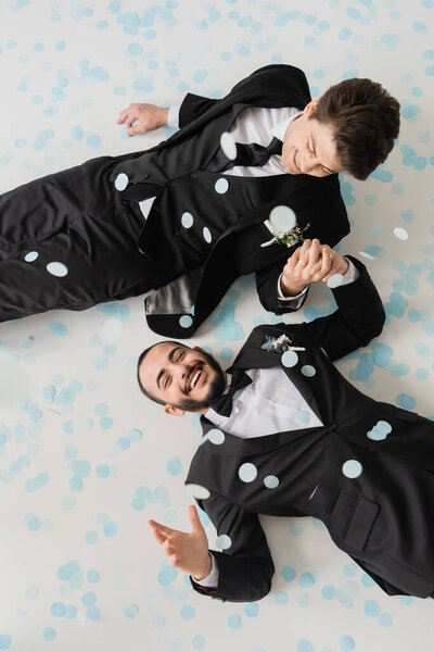 Top view of cheerful same sex couple in classic suits holding hands while having fun and lying on confetti during wedding celebration on grey background 