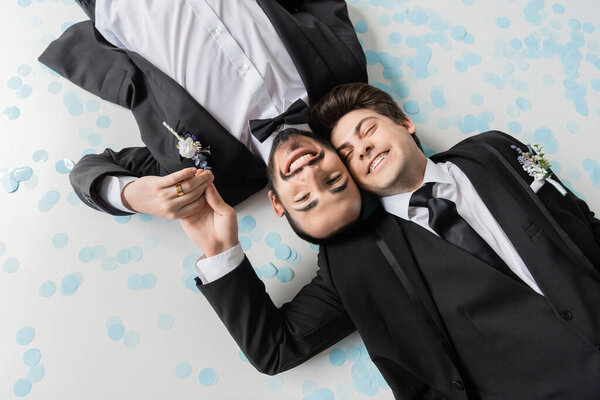 Top view of carefree same sex grooms in elegant suits holding hands and looking at camera while lying together on confetti on grey background 