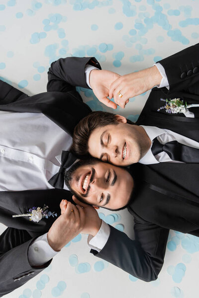 Top view of cheerful same sex couple in suits holding hands while lying together on confetti and smiling during wedding celebration on grey background 