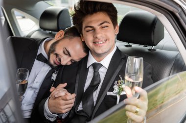 Smiling and young gay groom with braces in elegant suit with boutonniere holding glass of champagne and hand of boyfriend while sitting on backseat of car during honeymoon  clipart