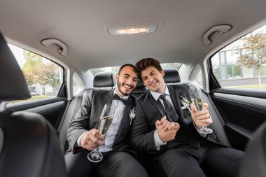 Düğünü arabanın arka koltuğunda kutlarken yakalarında yaka çiçekleri ve şampanya kadehleriyle gülümseyen homoseksüel çift. 