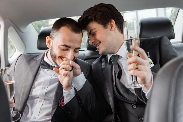 積極的なゲイ新郎でエレガントなフォーマル摩耗接吻手の若いパートナーでブレースと保持シャンパンガラスで新婚旅行中と座っている車の後部座席 — ストック写真