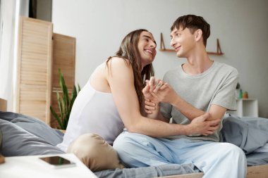 Gülümseyen ve genç eşcinsel çift pijama giyip, el ele tutuşup sohbet ederken evde komodinin yanında boş ekranlı, bulanık akıllı telefonun yanında oturuyorlar. 