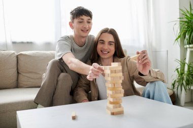 Genç ve mutlu homoseksüel çift, gündelik giysiler içinde gülümserken masada tahta bloklar oynuyorlar ve modern apartmanda birlikte vakit geçiriyorlar. 