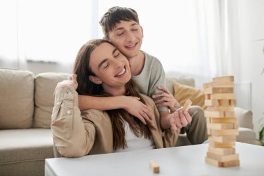 Young smiling gay man with closed eyes hugging long haired boyfriend in casual clothes near blurred wood blocks game on table in living room at home  clipart
