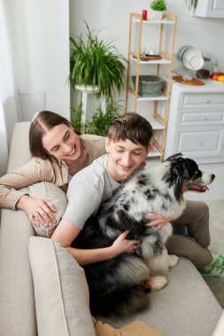Genç homoseksüel adam, evdeki oturma odasında kanepede dinlenirken gülümseyen uzun saçlı erkek arkadaşının yanında tüylü Avustralya çoban köpeğine sarılıyor. 
