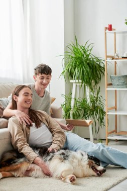 Neşeli eşcinsel adam kitap tutuyor ve rahat koltukta dinleniyor. Modern apartmandaki Avustralya çoban köpeğinin yanında uzun saçlı erkek arkadaşına sarılıyor. 