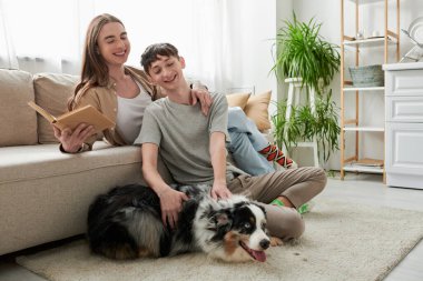 Neşeli eşcinsel adam halının üzerinde oturuyor ve Avustralya çoban köpeğine sarılıyor. Uzun saçlı genç partner, kitap tutuyor ve modern oturma odasındaki kanepede dinleniyor. 