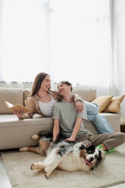 Neşeli eşcinsel bir adam halının üzerinde oturuyor ve Avustralyalı çoban köpeğine sarılıyor ve modern oturma odasında uzun saçlı, uzun saçlı bir partnere bakıyor. 