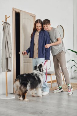 Modern koridorda palto askısının yanında duran tasmalı ve evde birlikte gülümseyen Avustralyalı çoban köpeğine bakan mutlu eşcinsel çift. 